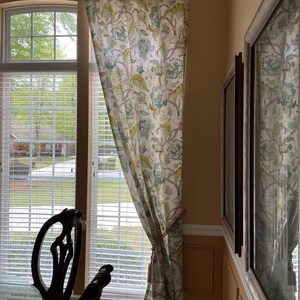 May include: A white curtain with a floral pattern in shades of green, blue, and yellow hangs in a window. The curtain is tied back with a rope.