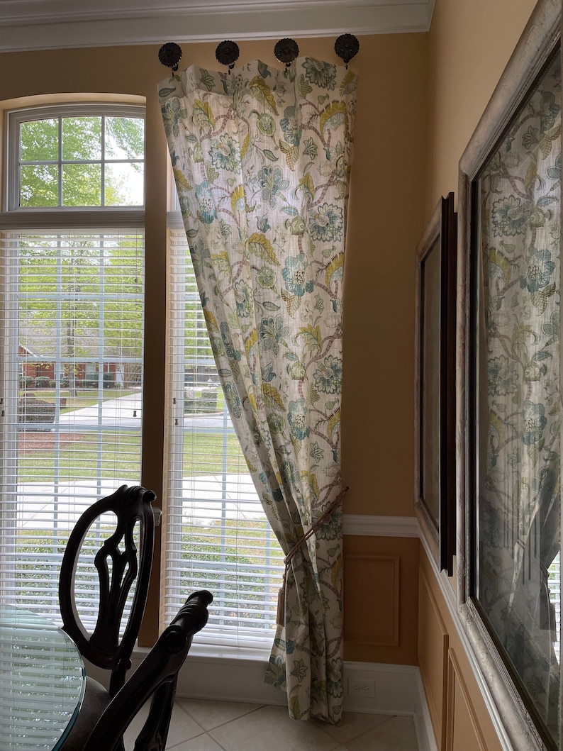 May include: A white curtain with a floral pattern in shades of green, blue, and yellow hangs in a window. The curtain is tied back with a rope.