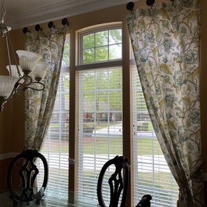 May include: A dining room with three windows featuring white blinds and floral patterned curtains. Two black chairs are positioned in front of the windows, with a glass table in the foreground.
