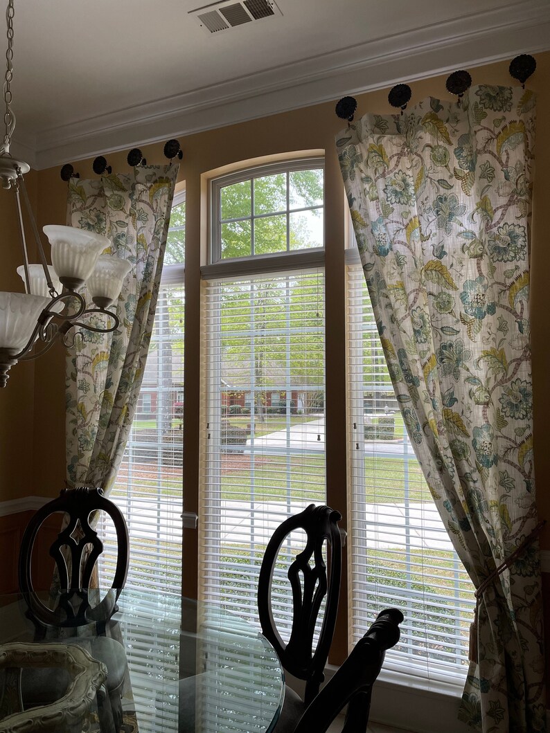 May include: A dining room with three windows featuring white blinds and floral patterned curtains. Two black chairs are positioned in front of the windows, with a glass table in the foreground.