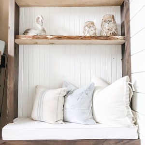 May include: A wooden shelf unit with a white cushion bench and a wood-burning fireplace below. The shelves are decorated with white ceramic swans, glass jars with rope accents, and driftwood.