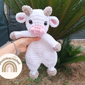 May include: A white crocheted plush toy cow with brown ears and a pink nose. The toy is being held by a hand.