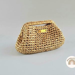 Può includere: Una borsa all'uncinetto color oro con una struttura rigida e un manico superiore. La borsa presenta un motivo intrecciato testurizzato e una piccola targhetta rettangolare in metallo dorato sul davanti con il testo plushikacrochet inciso.