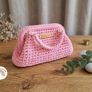 Può includere: Una borsa all'uncinetto rosa chiaro con una struttura rigida, un manico superiore singolo e una piccola placca metallica rettangolare color oro sul davanti. La borsa è realizzata in filato spesso con un motivo a maglia visibile e dettagli dorati.