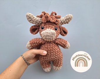 May include: A brown and beige crocheted plush toy in the shape of a Highland cow. The toy has a fluffy brown body, beige ears, and a beige face with black eyes. The toy is being held by a hand.