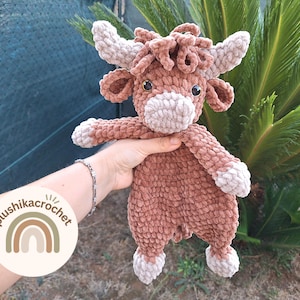 May include: A brown crocheted stuffed animal shaped like a cow with a white face and hooves. The cow is being held by a hand with a bracelet on the wrist. The background is a green plant.