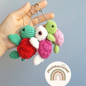 May include: Three crocheted turtle keychains with different coloured shells: red, pink, and green. Each turtle has a black eye and a green head. The keychains are attached to silver key rings. The image also includes a logo for the shop, "plushikacrochet", with a rainbow design.