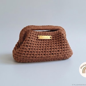 Può includere: Una borsa all'uncinetto marrone con una struttura rigida e una placca metallica rettangolare color oro sulla parte anteriore. La placca presenta il testo inciso Plushikacrochet. La borsa ha una costruzione in filato spesso e testurizzato.