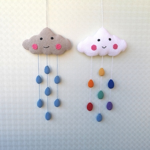 Könnte beinhalten: Zwei gehäkelte Wolken-Hängeschmuckstücke mit lächelnden Gesichtern. Eine Wolke ist beige mit blauen Regentropfen, die daran hängen, und die andere Wolke ist weiß mit regenbogenfarbenen Regentropfen, die daran hängen.