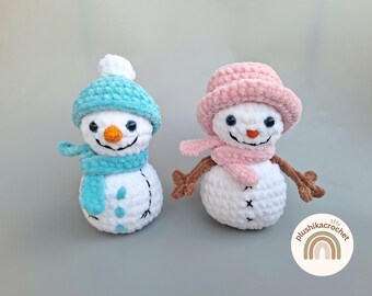 Op de afbeelding: Twee gehaakte sneeuwpop-versieringen, een met een blauwe muts en sjaal en de andere met een roze muts en sjaal. Beide sneeuwpoppen hebben een wit lichaam en zwarte ogen en mond.