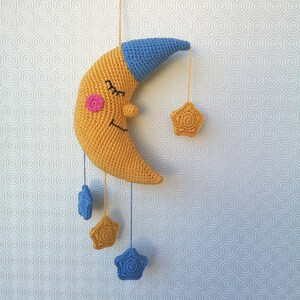 Könnte beinhalten: Eine gehäkelte gelbe Mondsichel mit einer blauen Schlafkappe und einem rosa Rouge. Der Mond hängt an einer Schnur mit drei gehäkelten blauen und gelben Sternen.