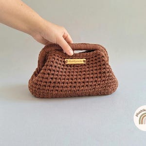 Puede incluir: Un bolso de mano de ganchillo marrón con una abertura de marco estructurado. El bolso presenta una construcción de hilo anudado texturizado y una pequeña etiqueta rectangular de madera centrada en el frente con el texto plushikacrochet grabado.
