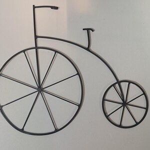 May include: Black metal bicycle wall art. The artwork depicts a penny-farthing bicycle with a large front wheel and a smaller rear wheel. The frame is curved, and the handlebars are straight with a curved top. The spokes are visible.