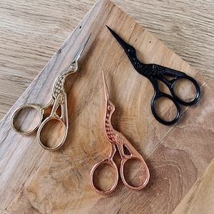 Puede incluir: Tres pares de tijeras decorativas con mangos en forma de pájaro. Un par es dorado, otro es de cobre y el último es negro.