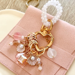 Puede incluir: Un llavero dorado en forma de corazón con un lazo de cuentas blancas. El llavero presenta varios dijes, que incluyen cuentas rosas y transparentes, colgantes en forma de concha y una gema rosa grande. El fondo incluye una bolsa rosa y unas tijeras doradas.