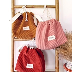 Op de afbeelding: Drie corduroy trekkoordtassen in tinten roest, roze en rood hangen aan een bamboeladder. Elke tas heeft een witte band en een klein rechthoekig label met de tekst "BARBARA".