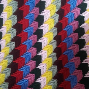 May include: Close-up of a colorful, textured textile with a repeating geometric pattern. The design features rows of stacked blocks in various colors, including red, blue, yellow, pink, and green, set against a dark background. The fabric appears to be knitted or crocheted.