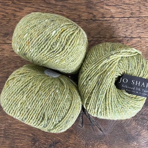 以下が含まれることがあります： 黄色と茶色の斑点のある緑色の糸のボールが3つ。糸には「Silkroad DK Tweed 45% Wool 10% Silk 5% Cashmere」と「JO SHARP」のラベルが付いています