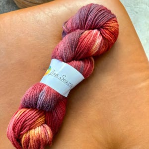 May include: A skein of hand-dyed yarn in shades of red, orange, and purple. The yarn is wrapped with a white label that reads "Lisa Souza Knitwear and Dyeing". The yarn is resting on a brown leather surface.
