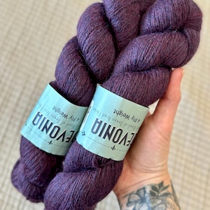 May include: Two skeins of deep purple yarn are held in a hand. The yarn is labeled "EVONIA" and "4 Ply Weight". The yarn has a soft, textured appearance. The background is a light beige color.