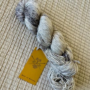 May include: A ball of speckled yarn in shades of grey and dark blue, with a yellow tag attached. The tag features a logo and the text "Farmers Daughter Fibers" and "Made in Montana". The yarn is displayed on a textured, neutral-coloured surface.