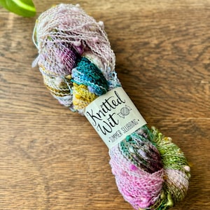 May include: A skein of colorful yarn with a label that reads "Knitted Wit Summer Slubbing." The yarn features a blend of pink, green, blue, and yellow hues. The yarn is displayed on a wooden surface.