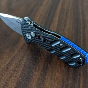 Puede incluir: Un cuchillo plegable negro con un mango texturizado y un acento azul. La hoja está parcialmente abierta y tiene un acabado plateado.