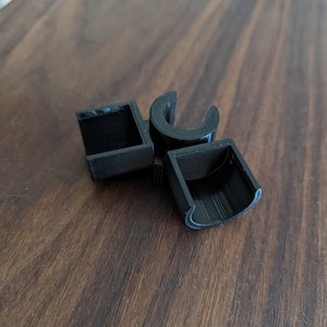 May include: Black plastic 3D printed object with a square and two curved shapes. The object is on a wooden surface.