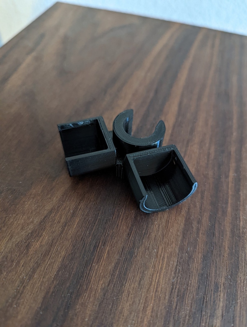May include: Black plastic 3D printed object with a square and two curved shapes. The object is on a wooden surface.