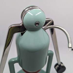 Pode incluir: Uma cafeteira azul claro e prateada com uma mangueira longa e flex&iacute;vel. A cafeteira tem uma parte superior redonda e bulbosa e uma base com um padr&atilde;o de grade.