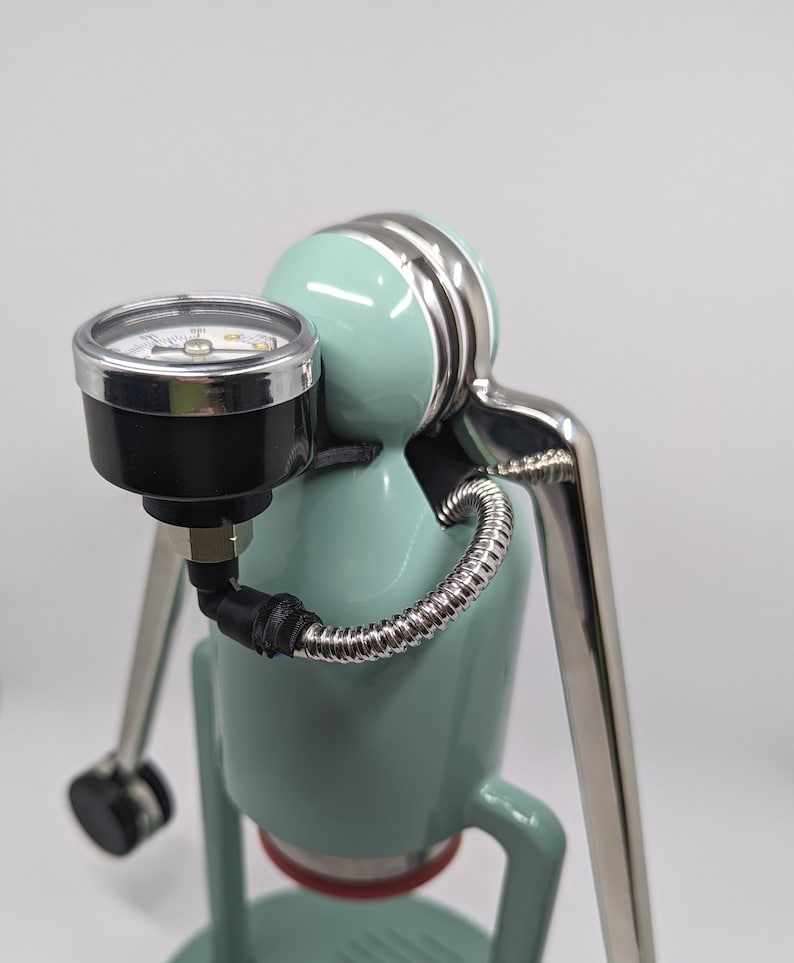 Pode incluir: Uma m&aacute;quina de caf&eacute; espresso verde menta com uma alavanca prateada e um man&ocirc;metro. O man&ocirc;metro est&aacute; conectado a uma mangueira preta que envolve a m&aacute;quina.