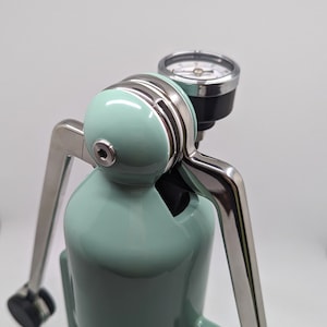 Pode incluir: Uma m&aacute;quina de caf&eacute; espresso manual de cor verde menta com uma alavanca prateada e um man&ocirc;metro. A m&aacute;quina tem a palavra "Cafelat" impressa na frente.