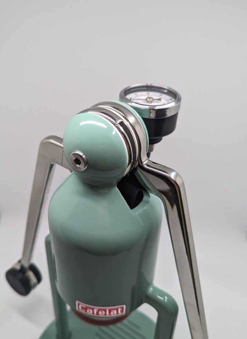 Pode incluir: Uma m&aacute;quina de caf&eacute; espresso manual de cor verde menta com uma alavanca prateada e um man&ocirc;metro. A m&aacute;quina tem a palavra "Cafelat" impressa na frente.