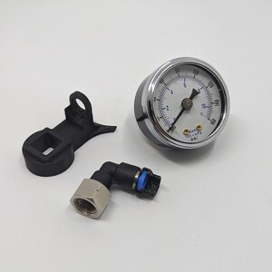 Pode incluir: Um man&ocirc;metro prateado com um mostrador branco e marcas pretas. O man&ocirc;metro l&ecirc; at&eacute; 180 PSI e 10 BAR. O man&ocirc;metro est&aacute; conectado a um encaixe de pl&aacute;stico preto com uma mangueira preta e azul.