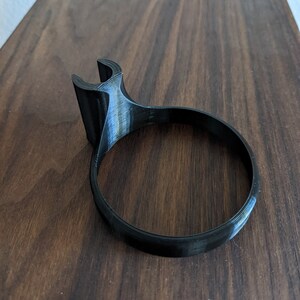 May include: A black plastic 3D printed phone stand with a curved base and a hook on the side.