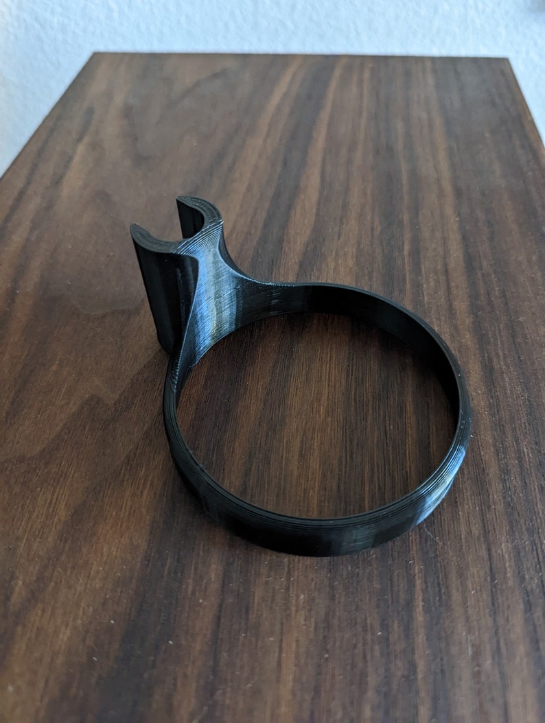 May include: A black plastic 3D printed phone stand with a curved base and a hook on the side.