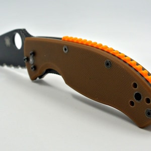 Puede incluir: Un cuchillo plegable marrón con una hoja negra y un mango texturizado naranja.