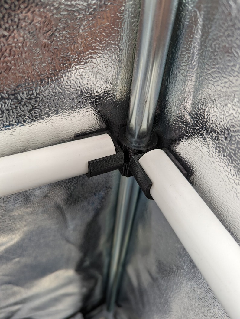 May include: Close-up of a black plastic connector joining three white pipes to a silver metal pole. The connector is used to assemble a grow tent frame.