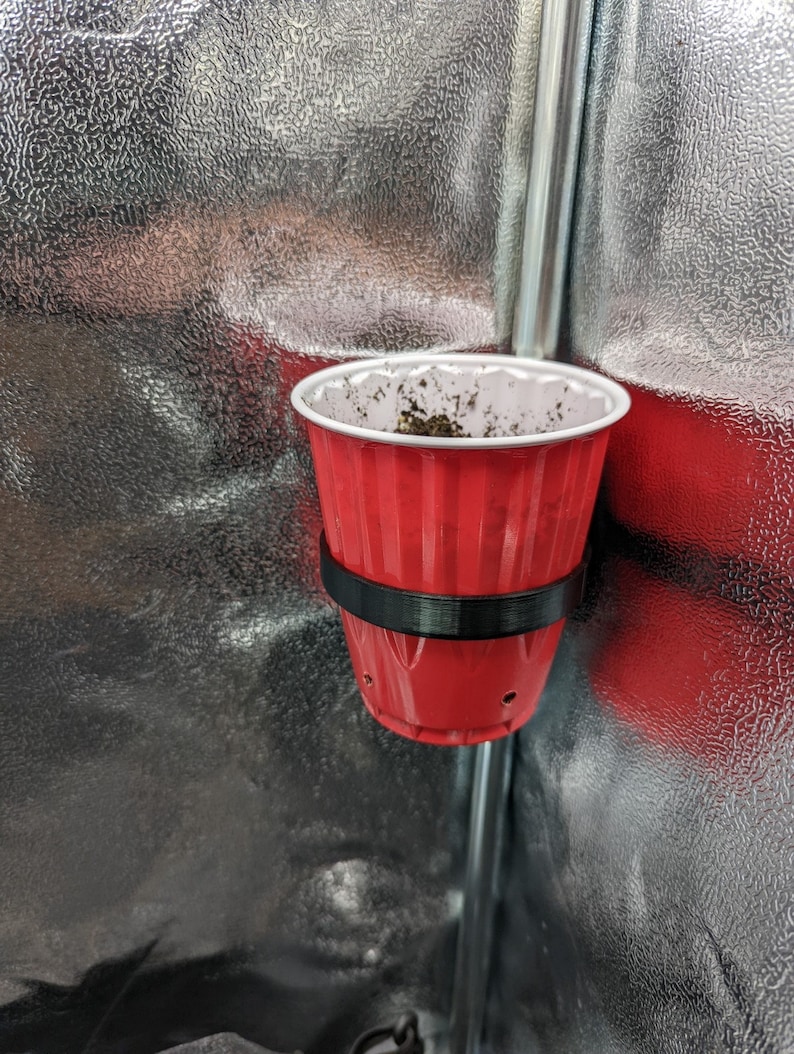 May include: A red plastic cup with drainage holes is attached to a metal pole with a black plastic band. The cup is filled with soil.