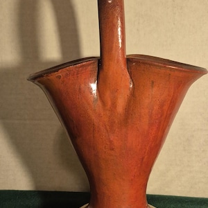 Könnte beinhalten: Ein handgefertigter Keramikkorb mit leuchtend orange-roter Glasur. Der Korb hat einen ausgestellten oberen Rand, einen stabilen Griff und einen gelben Sockel. Das Design erinnert an Vintage-Keramik.