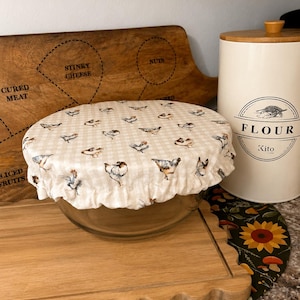 May include: A round fabric bowl cover with an elasticated ruffled edge, featuring a beige and white gingham pattern decorated with small illustrations of chickens. The cover is placed over a clear glass bowl, creating a snug fit for storage.
