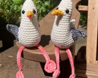 Op de afbeelding: Twee handgemaakte gehaakte zeemeeuwfiguren. De zeemeeuwen hebben witte lichamen, grijze vleugels en gele snavels. Hun poten zijn roze en rood en ze hebben zwarte ogen en rode snavels. Ze zitten op een houten oppervlak.