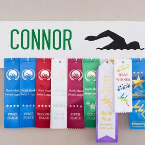 May include: A white wooden sign with the name "CONNOR" in green letters and a black swimmer silhouette. Below, ribbons in various colors display "FIRST PLACE", "SECOND PLACE", "FIFTH PLACE", and "Eighth Place" ribbons, with "HEAT WINNER" ribbons.