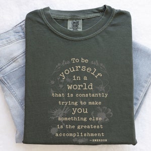May include: A dark green t-shirt with a white floral design and the text "To be yourself in a world that is constantly trying to make you something else is the greatest accomplishment - EMERSON".