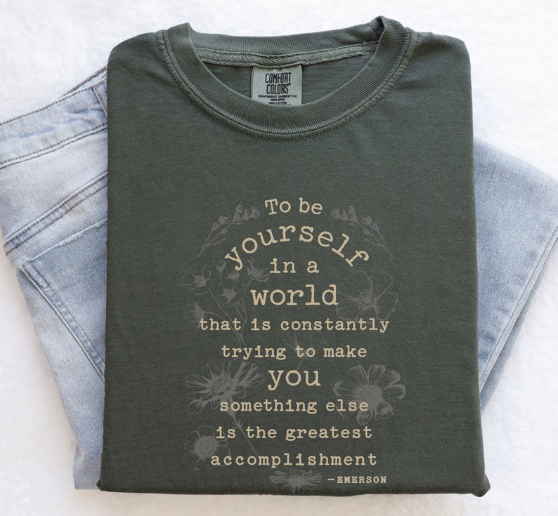 May include: A dark green t-shirt with a white floral design and the text "To be yourself in a world that is constantly trying to make you something else is the greatest accomplishment - EMERSON".