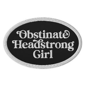 May include: Oval-shaped embroidered patch with the words "Obstinate Headstrong Girl" in white script on a black background. The patch has a silver-colored border.