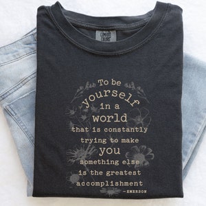 May include: A dark gray t-shirt with a white graphic print that reads "To be yourself in a world that is constantly trying to make you something else is the greatest accomplishment - EMERSON".