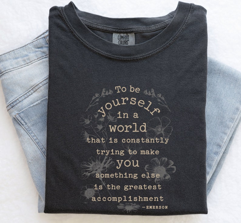 May include: A dark gray t-shirt with a white graphic print that reads "To be yourself in a world that is constantly trying to make you something else is the greatest accomplishment - EMERSON".