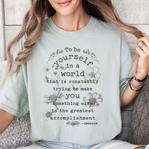 May include: A light blue t-shirt with a floral design and the quote "To be yourself in a world that is constantly trying to make you something else is the greatest accomplishment - EMERSON".