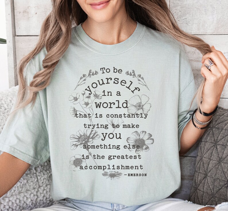 May include: A light blue t-shirt with a floral design and the quote "To be yourself in a world that is constantly trying to make you something else is the greatest accomplishment - EMERSON".